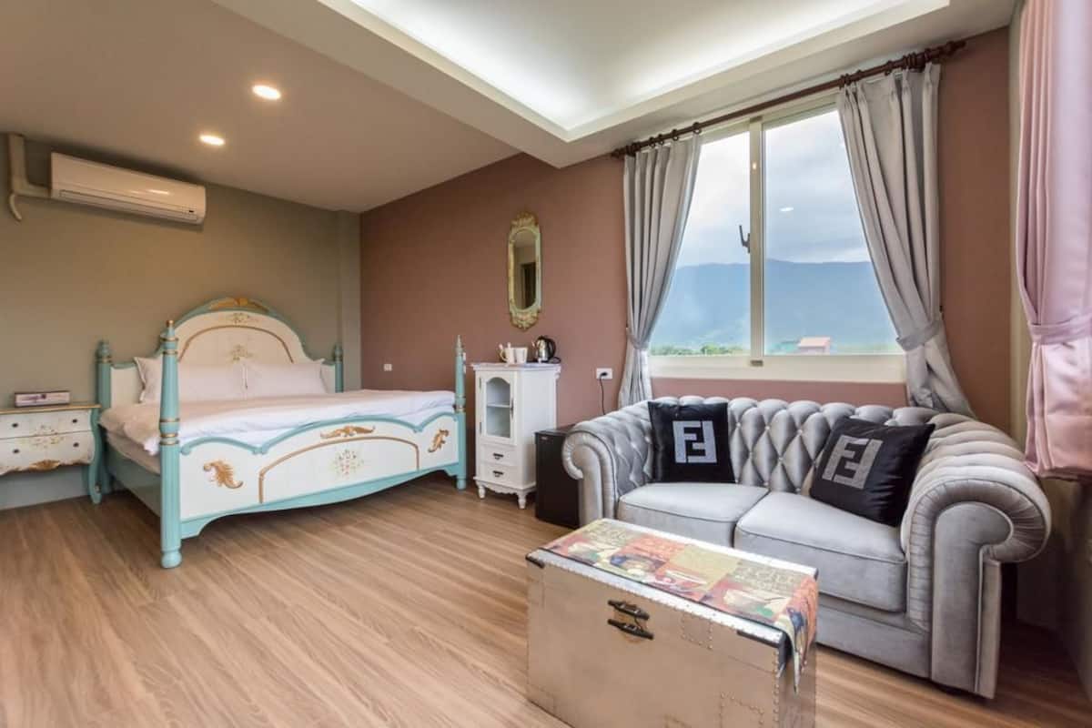 A bedroom with a bed, a dresser, a vanity, a sofa with pillows, a window with curtains, and a suitcase.