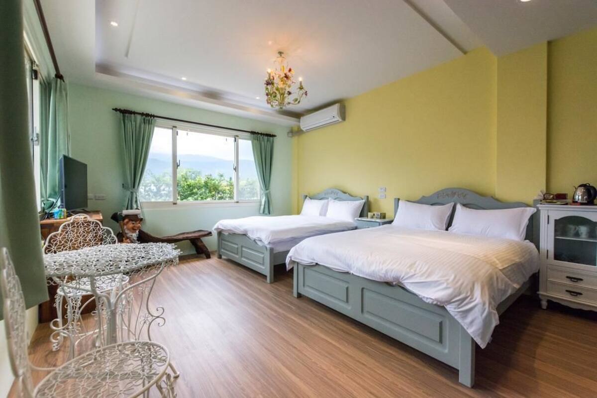 A bedroom with two beds, a chandelier, a TV, and a view of greenery outside.