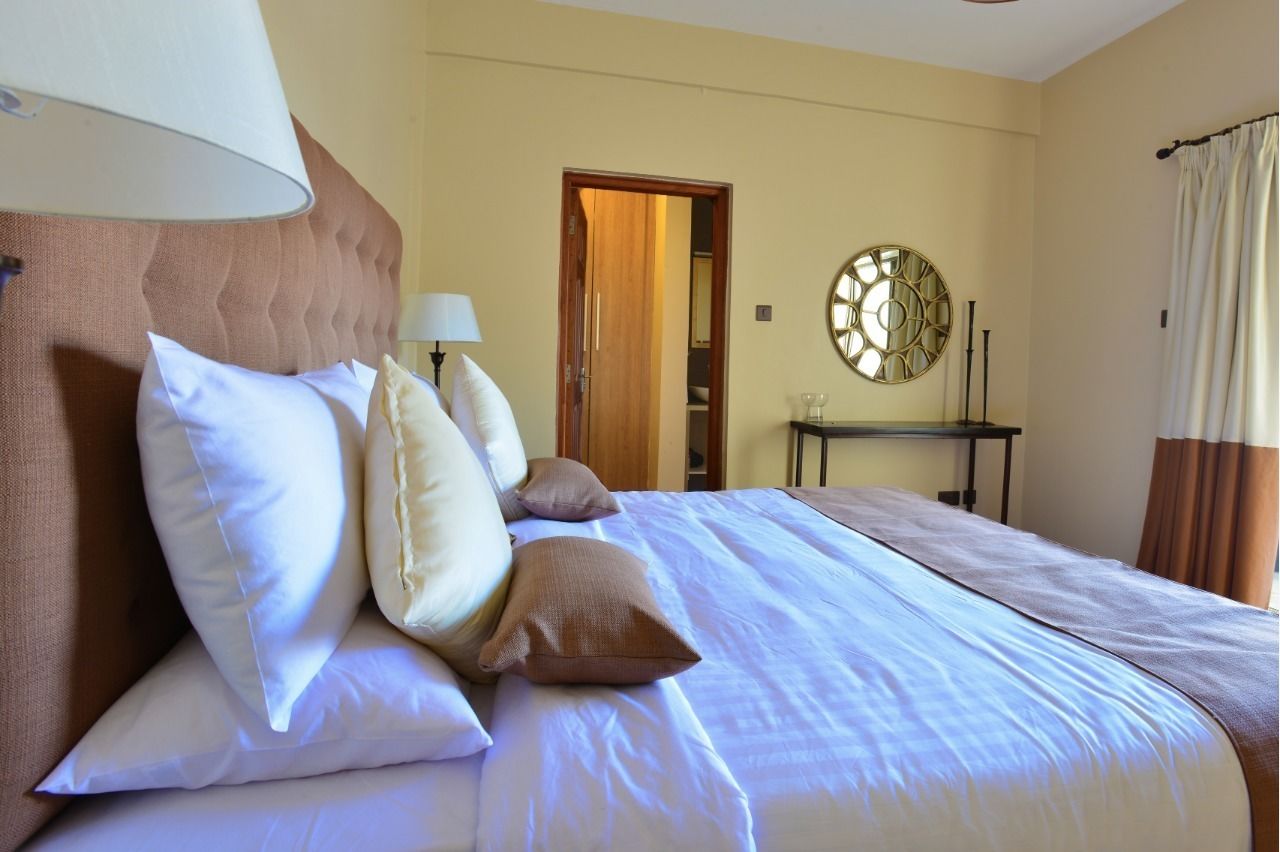A hotel room with a bed, pillows, a lamp, a mirror, and a door leading to another room.