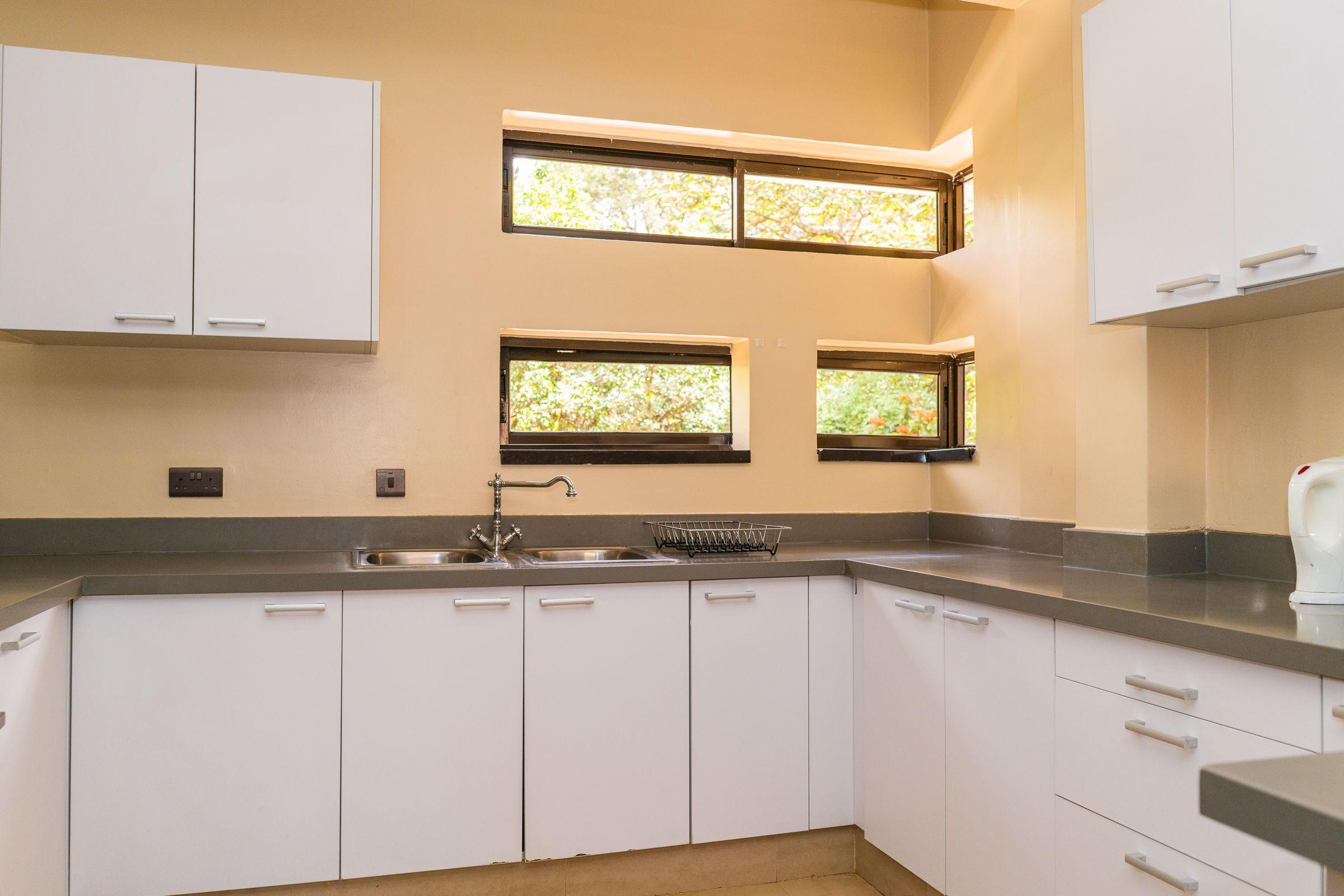 A modern kitchen with white cabinets, a stainless steel sink, and three windows with a view of greenery.