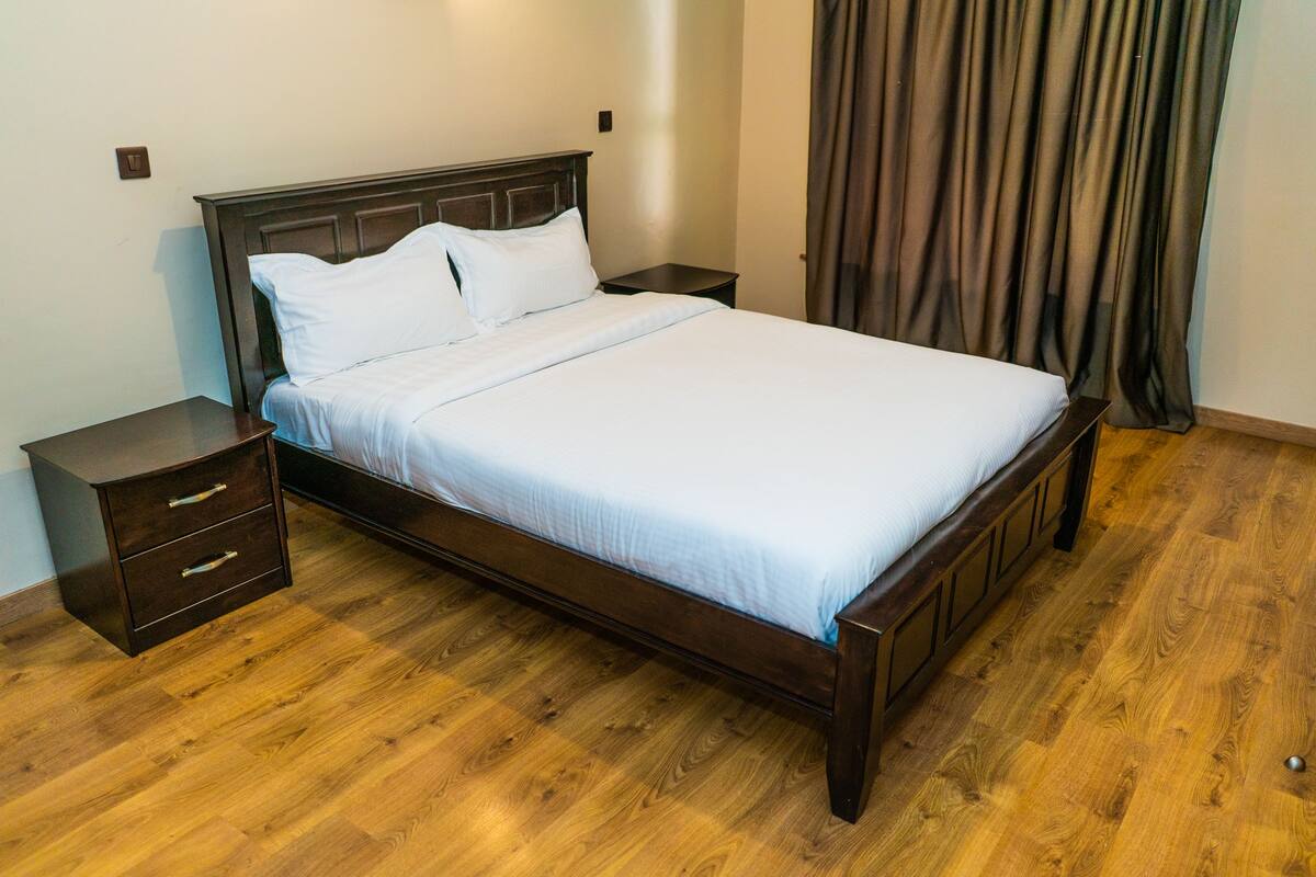 A neatly made bed with a wooden frame and white bedding in a room with wooden flooring and a dark-colored headboard.