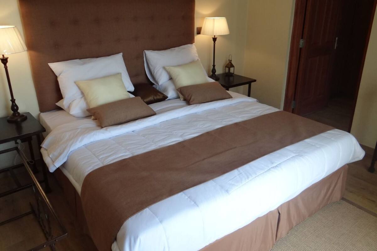 A neatly made bed with a brown headboard and white bedding, two bedside tables with lamps, and a wooden door in the background.