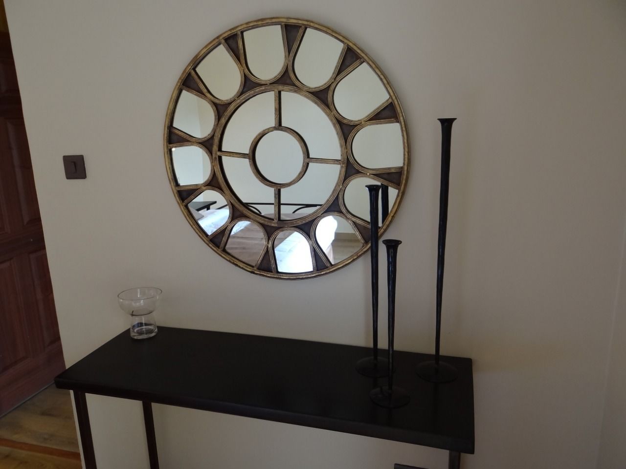 Un espejo redondo con un diseño geométrico en una pared, una mesa negra con un cuenco de vidrio y dos candelabros negros.