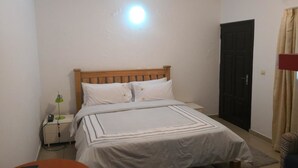 A bedroom with a wooden bed, white bedding, a nightstand with a lamp, a black door, and a window with curtains.