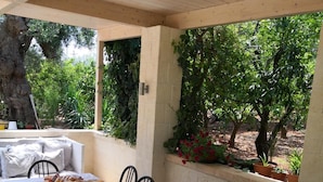 Uma área de refeições ao ar livre com uma mesa comprida posta para uma refeição, rodeada por cadeiras, com vista para árvores e plantas.