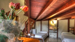 Quarto rústico com vigas de madeira, duas camas de solteiro, uma mesinha com uma cadeira e um vaso com flores.
