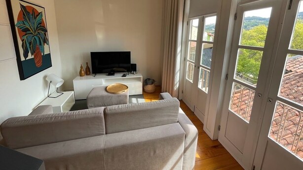 A modern living room with a sofa, a TV, and a dining table. There are large windows with a view of a red-tiled roof and greenery. - Mezzanine Apartment in the heart of Guimarães. (Guimarães)