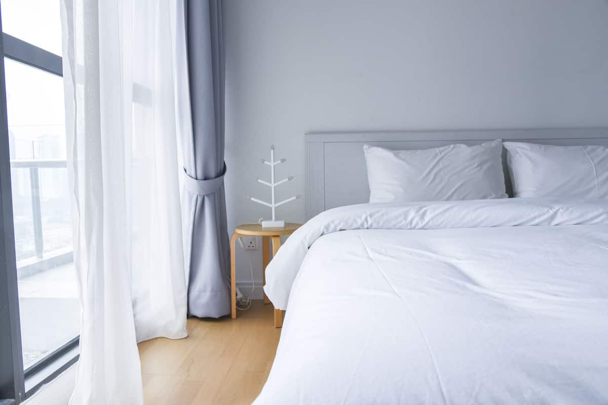 A modern hotel room with a large bed, a bedside table with a lamp, and a view of the city through the window.