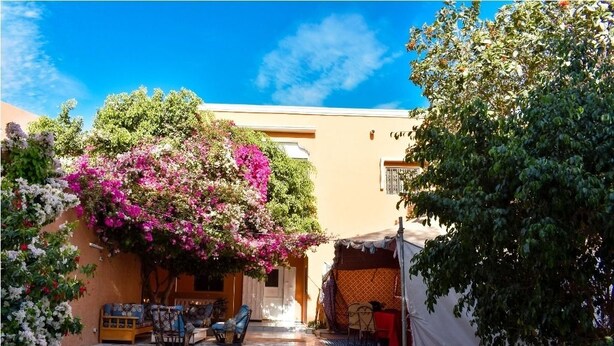 Un patio con una zona de estar cubierta, piso de baldosas y un edificio de fachada color crema.