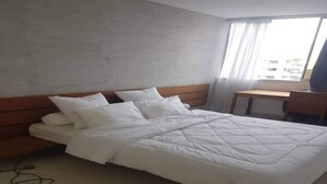 A neatly made bed with white bedding in a room with a wooden headboard, a small wooden desk, and a window with curtains. - Résidence Djény T3 (Abidjan)