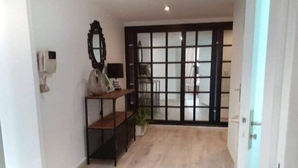 A hallway with a wooden floor, a black-framed mirror, a small table with a lamp, and a sliding glass door leading to another room. - Résidence Djény T3 (Abidjan)