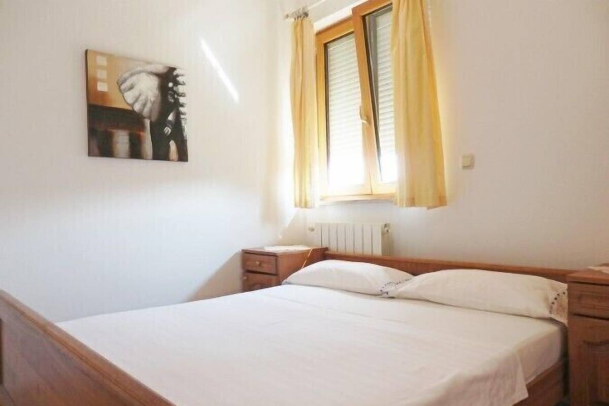 Quarto com uma cama de solteiro, mesa de cabeceira, janela com cortinas e um quadro na parede.