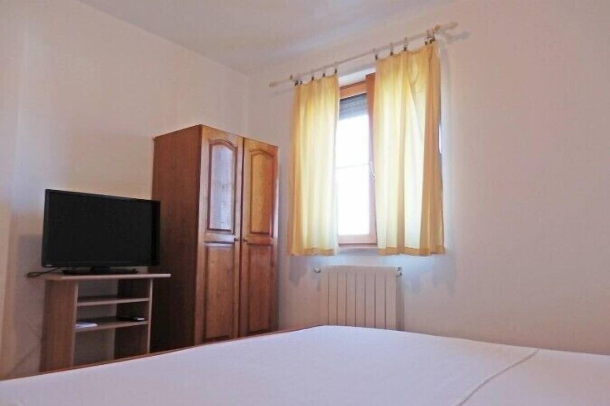 Quarto com cama, televisão sobre um suporte, armário e janela com cortinas.
