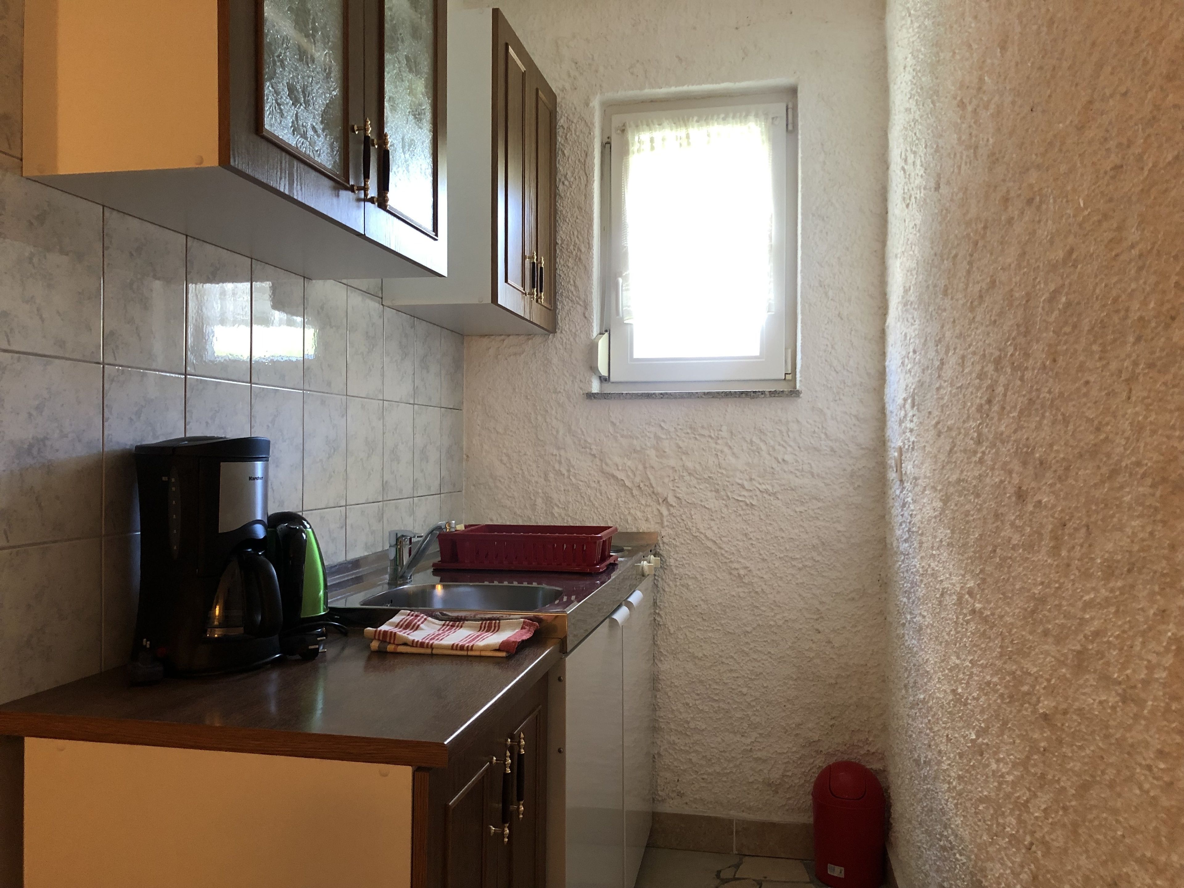 A compact kitchen with a coffee maker, kettle, and red dish rack. A window provides natural light, and a red fire extinguisher is visible.