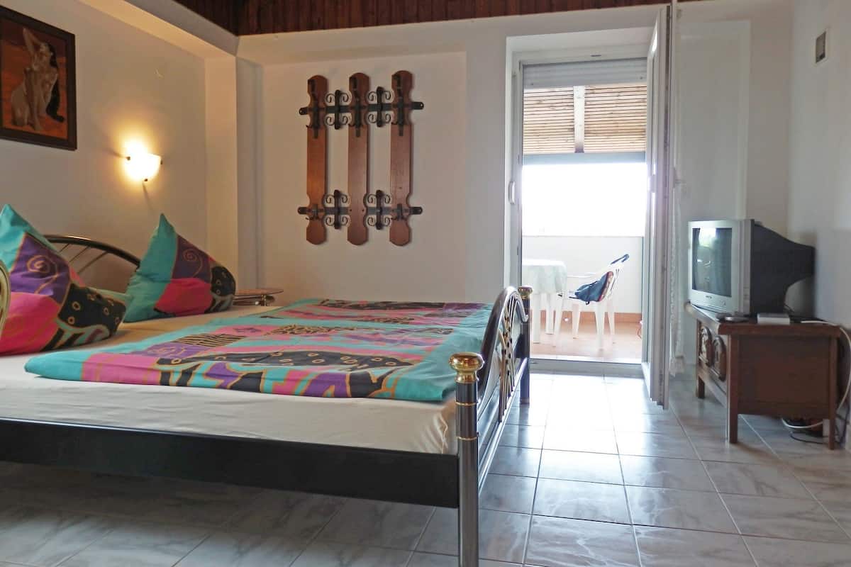 Un dormitorio con cama, almohadas coloridas, televisor montado en la pared y balcón con mesa y sillas.