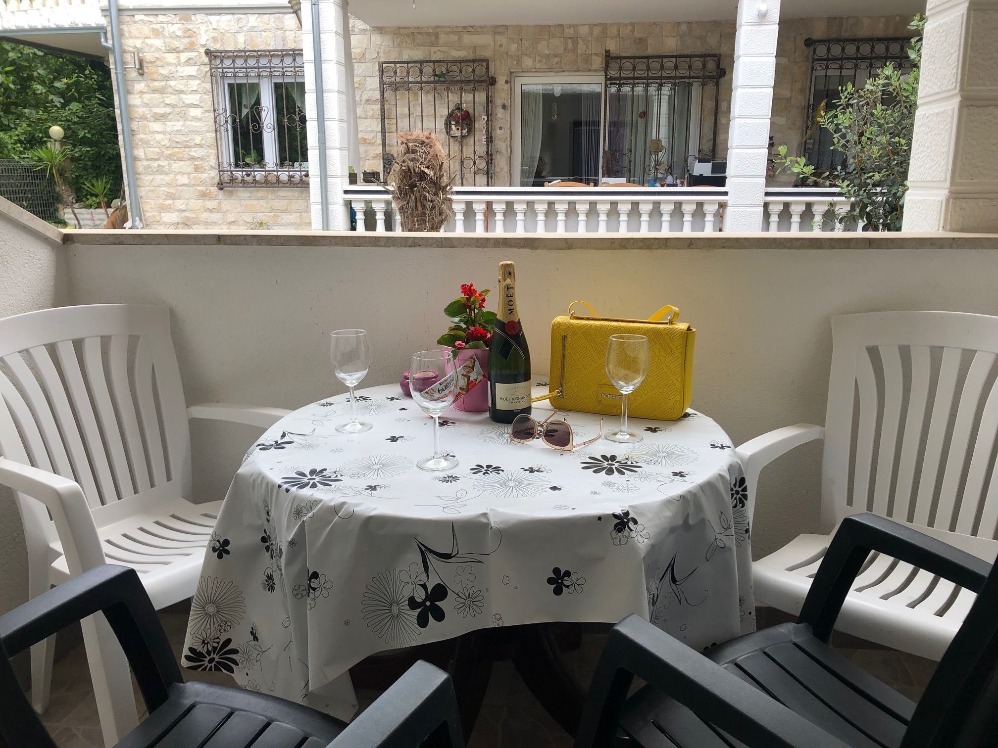 A round table with a floral tablecloth, two wine glasses, a bottle of Moët champagne, and a yellow handbag on a balcony with white chairs.