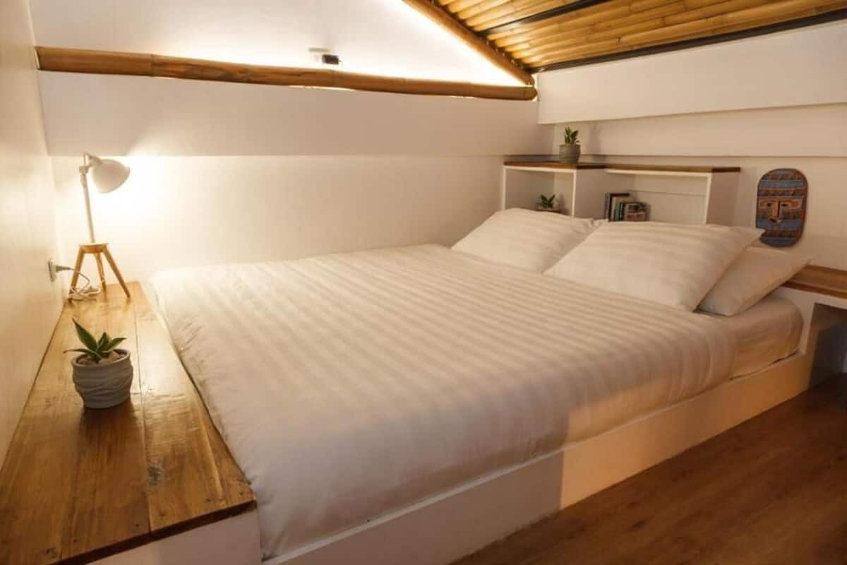 Ein Einzelbett mit Holz Kopfteil, ein Nachttisch mit Lampe sowie ein kleines Regal mit Büchern und Pflanze.