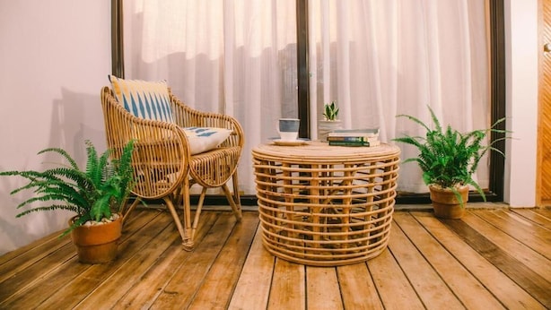 Une chaise en osier avec un coussin, une table ronde en osier et deux plantes en pot, le tout posé sur un plancher en bois.