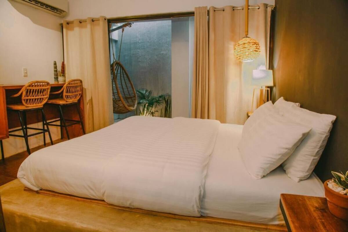 Ein Bett mit weißer Bettwäsche, eine hölzerne Bank, zwei Korbstühle, eine Hängelampe und ein Blick auf einen Balkon mit Vorhängen.