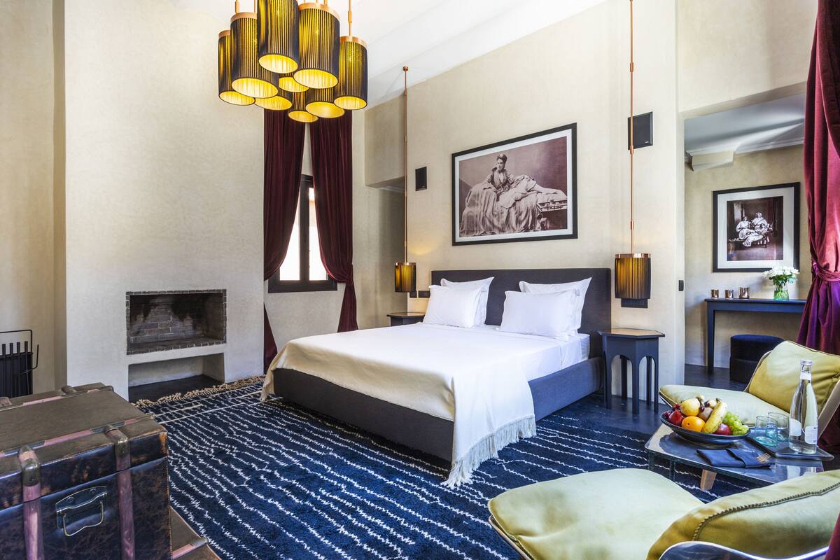 A hotel room with a large bed, a fireplace, a dining area with a table and chairs, and a large window with curtains.