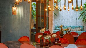 A dining area with a long table, multiple chairs, hanging pendant lights, and a view of a blue wall and a plant. - Riad Tajania (Marrakech)