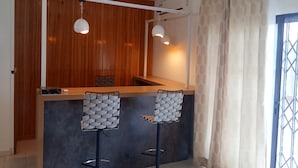 A modern kitchen with wooden paneling, a black countertop, two bar stools with woven seats, and pendant lighting. - Résidence Akwaba T7 (Abidjan)