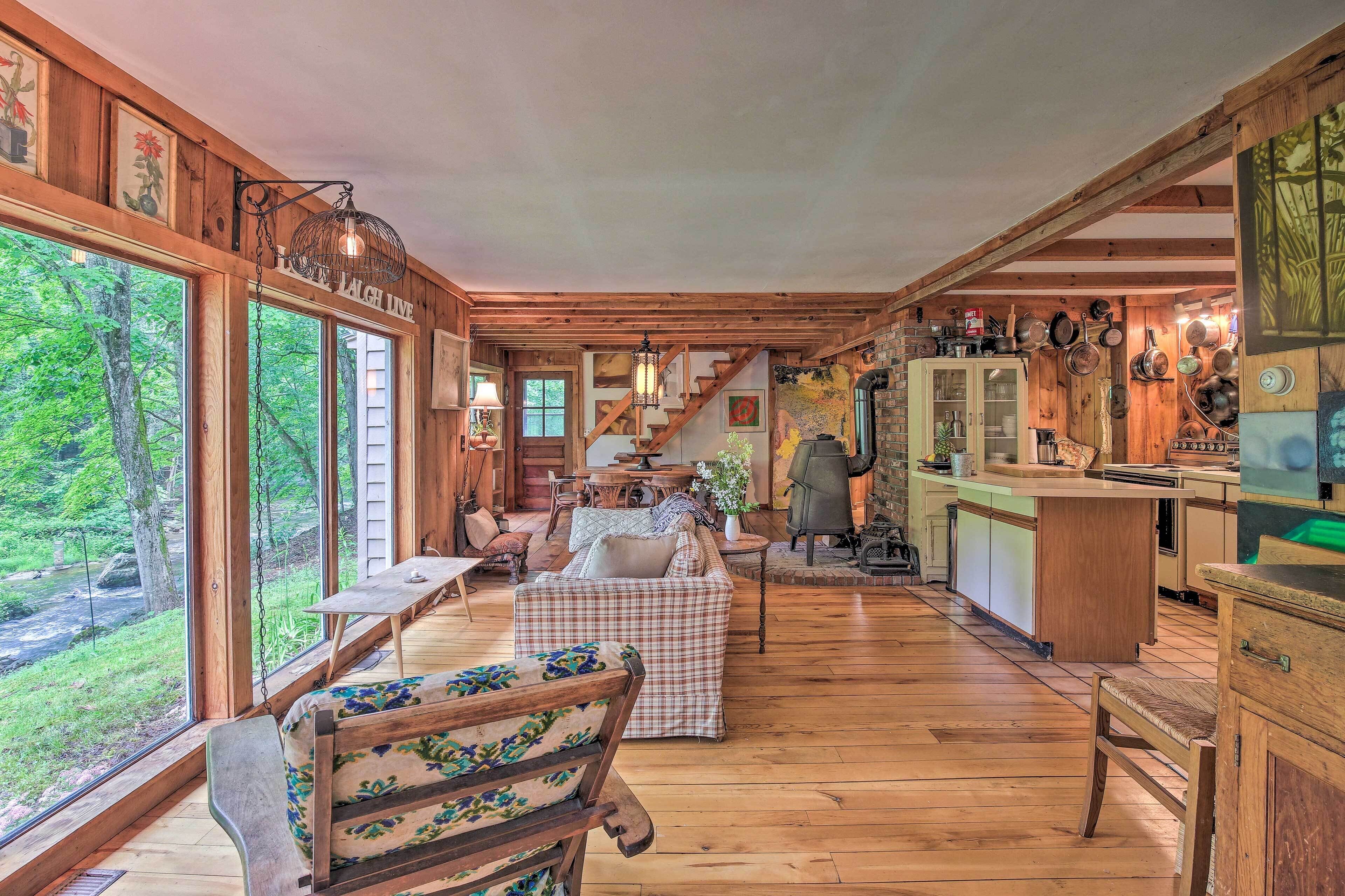 L’intérieur d’un chalet rustique, avec un plancher en bois, un coin repas avec un banc, une cuisine dotée de meubles en bois et de grandes fenêtres donnant sur une forêt.