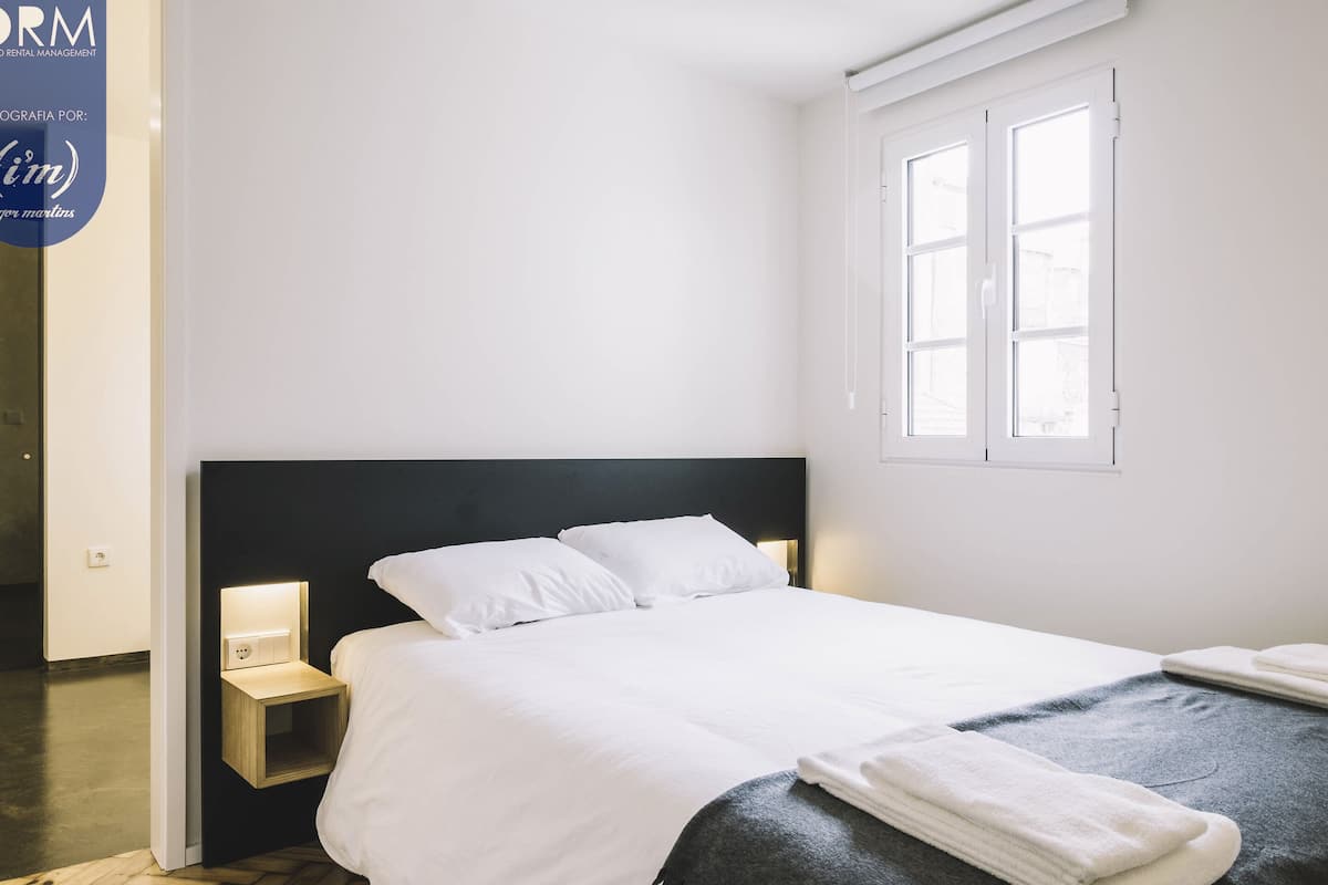 Uma cama bem arrumada com lençóis brancos, cabeceira escura e mesa de cabeceira com abajur.