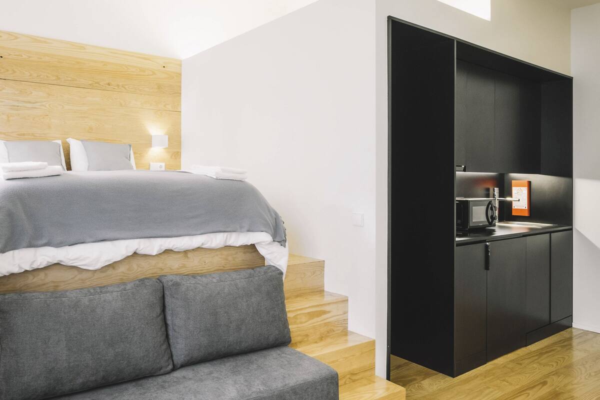 Quarto de hotel moderno com cama, sofá cinza, cabeceira de madeira, armário preto e uma pequena cozinha equipada.