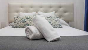 Un lit bien fait, avec une tête de lit capitonnée, deux coussins à motifs floraux et une serviette enroulée posée sur une couverture grise à texture.