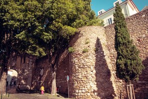 Eine steine Mauer mit einem schmalen Durchgang, ein hoher Baum und ein Gebäude im Hintergrund.