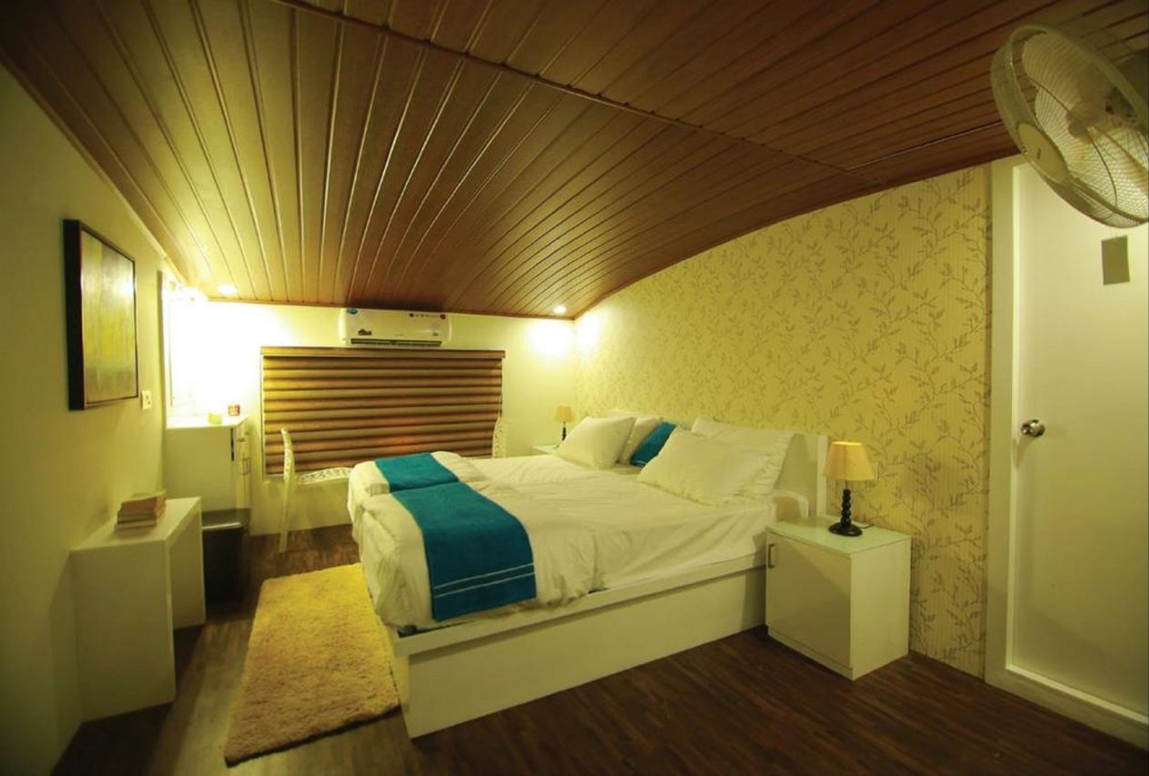 Un dormitorio con techo de madera, una cama con ropa de cama blanca y un plaid azul, mesitas de noche con lámparas y un aire acondicionado instalado en la pared.