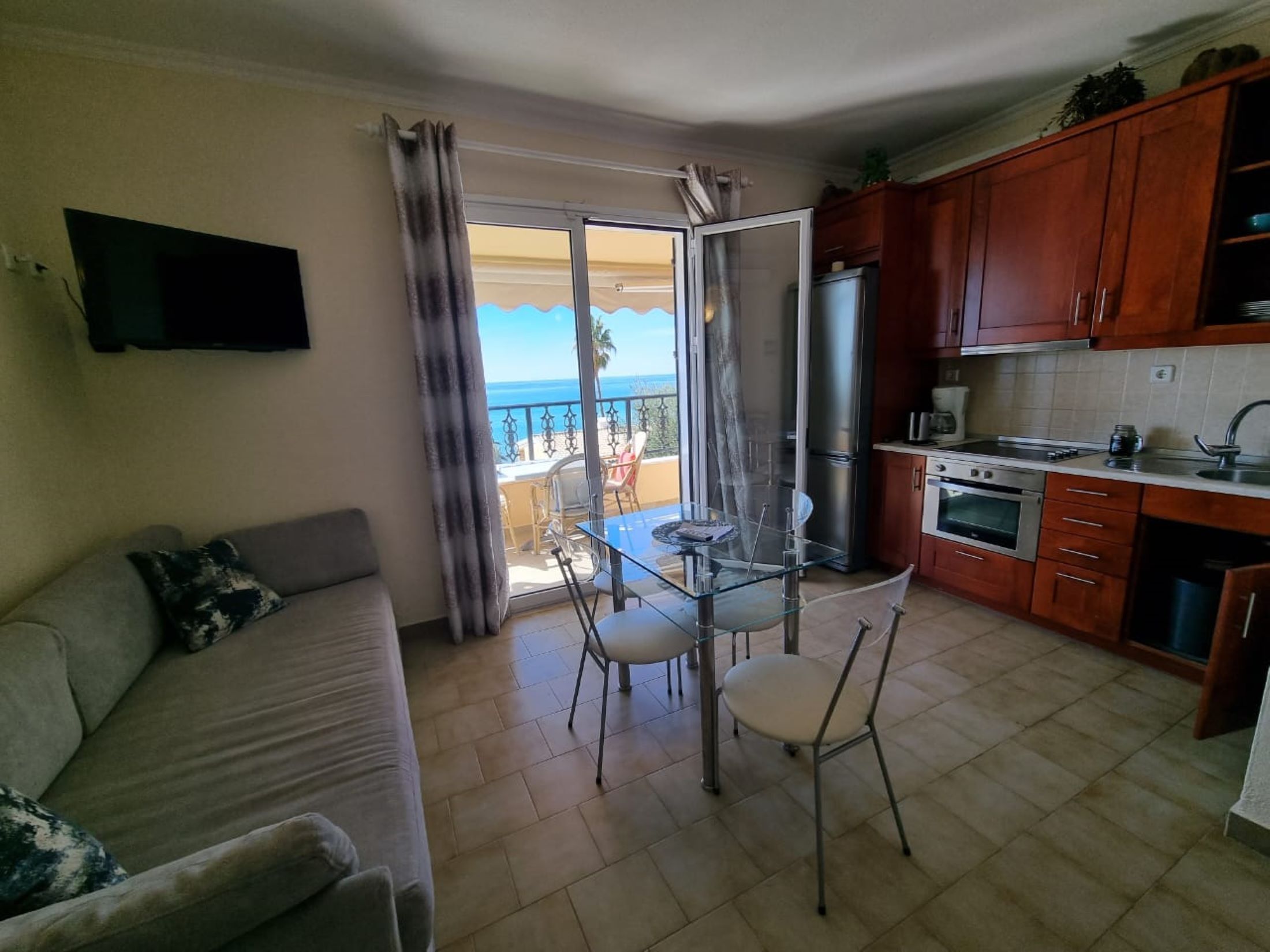 A living room with a sofa, a glass dining table with chairs, a kitchen with wooden cabinets, a refrigerator, and an oven. There is a sliding glass door leading to a balcony with a view of the sea.