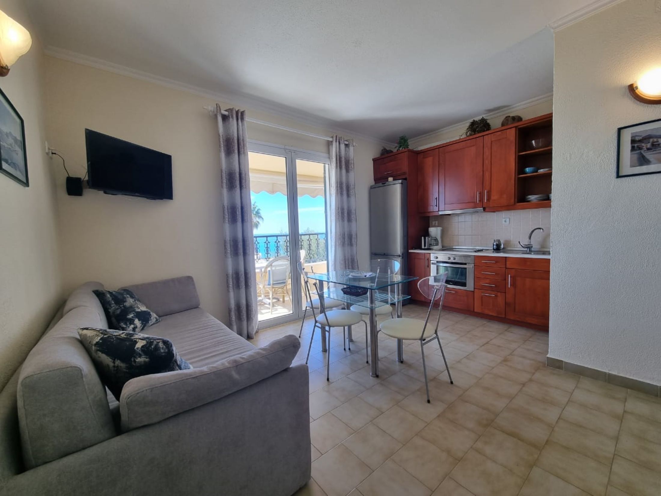 A living room with a sofa and a glass dining table, a kitchen with wooden cabinets, and a balcony with a view.