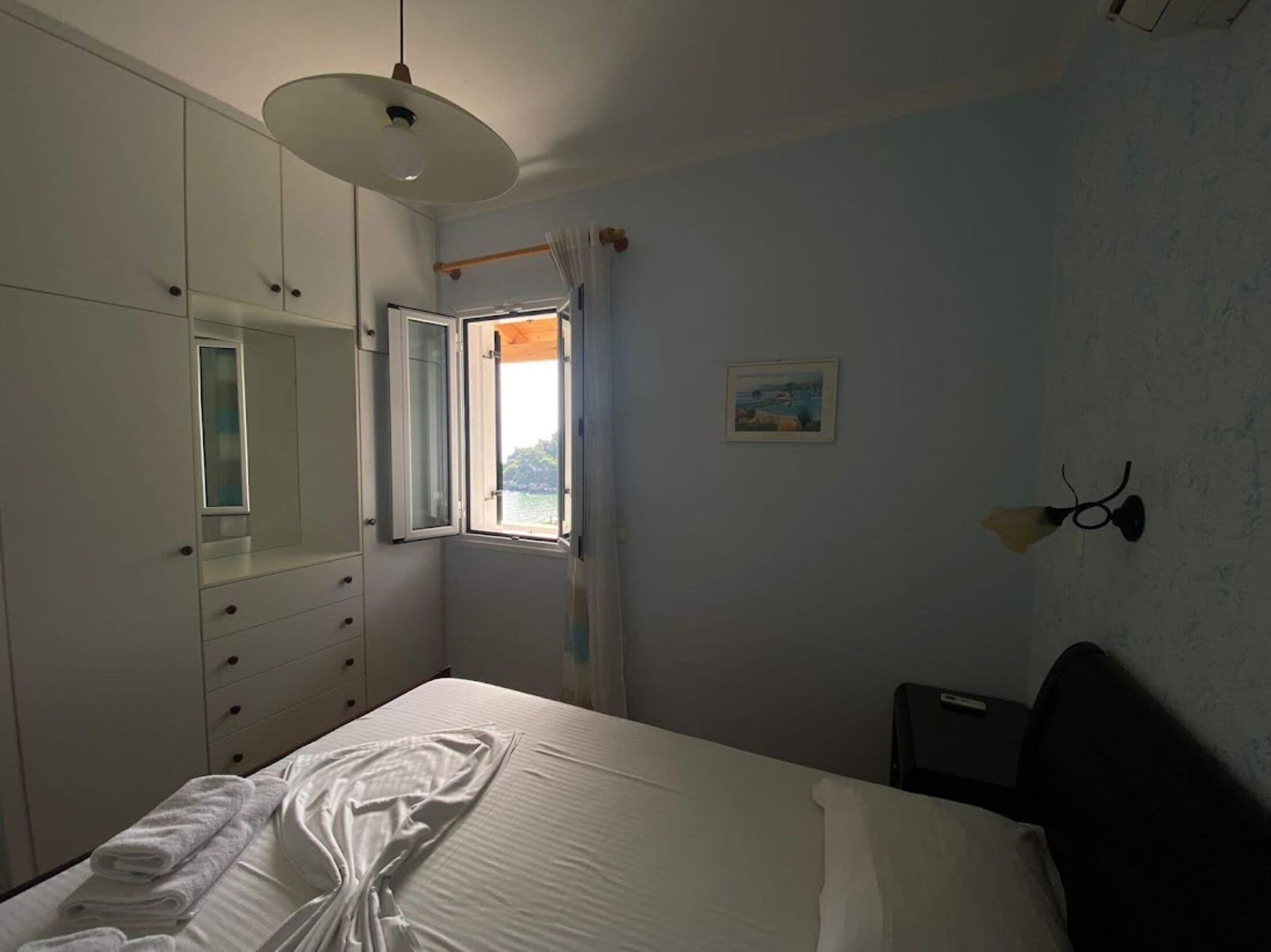 Un dormitorio con cama, armario, ventana con cortinas, un cuadro y una lámpara de pared.