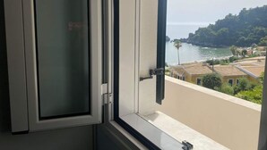 A window with a view of a coastal area, including a beach, buildings, and greenery. - Corfu Island Apartment 86 (Corfu)