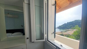 A room with a bed, a small cabinet, and a window with a view of a coastal landscape. - Corfu Island Apartment 86 (Corfu)
