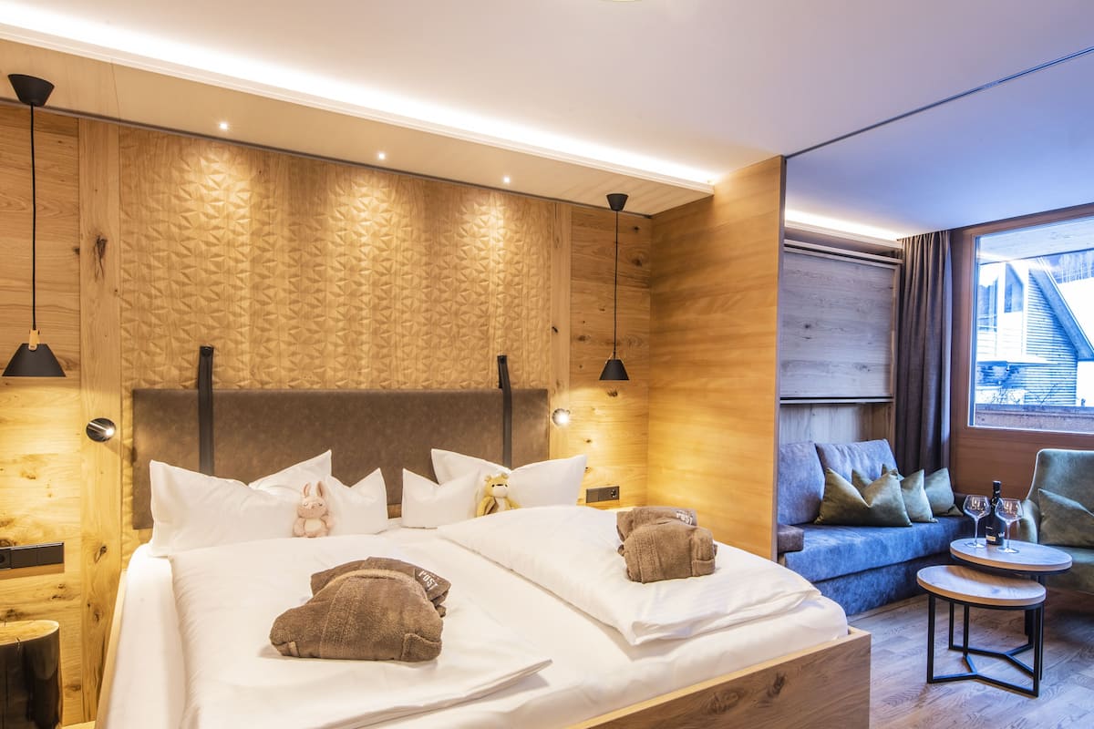 A modern hotel room with a wooden headboard, a sofa, a small table with a wine glass, and a teddy bear on the bed.