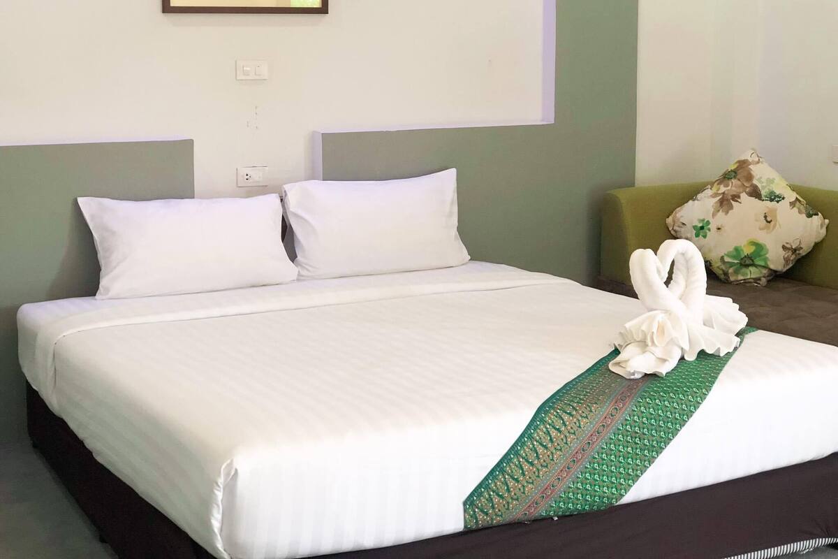 Una cama bien tendida con sábanas blancas, un lazo decorativo verde y dorado, y una toalla blanca doblada en forma de cisne.