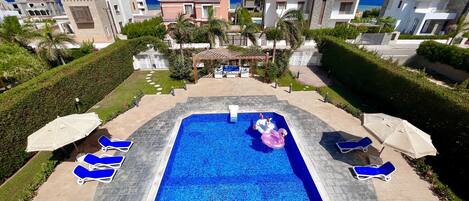 A swimming pool with blue tiles, surrounded by lounge chairs and umbrellas, with a view of the sea and modern buildings in the background.