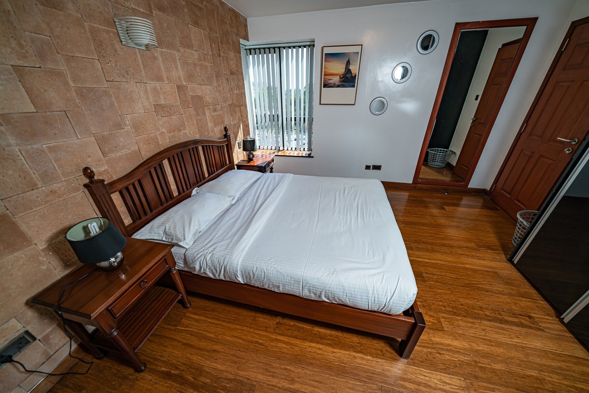 A bedroom with a wooden bed, a nightstand with a lamp, a window with blinds, a picture on the wall, and a door leading to another room.