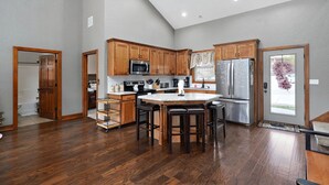 A modern kitchen with wooden cabinets, a central island with stools, stainless steel appliances, and a view through a glass door. - Pool&SplashPad Hot Tub PoolTable, Foosball, Lake, Shows, Golf, Shop, FUN (Hollister)