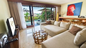 A living room with a sofa, a coffee table, and a TV. There is a window with a view of a pool and greenery. - Karma Luxury 2 bd. VILLA *TOP 5 stars BALi RESORT* (Ungasan)