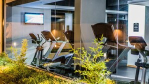 A gym with stationary bikes and a treadmill, glass walls, and a TV on the wall.