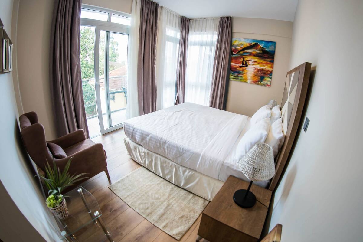 Een slaapkamer met een groot bed, een bureau, een stoel, een plant en een schilderij aan de muur.
