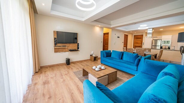 A modern living room with a large blue sofa, a coffee table with teapots, a flat-screen TV mounted on the wall, and a kitchen area in the background.