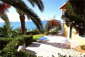 A stone patio with sun loungers and a small table, surrounded by lush greenery and a view of the sea. - Corfu Island Apartment 45 (Corfu)