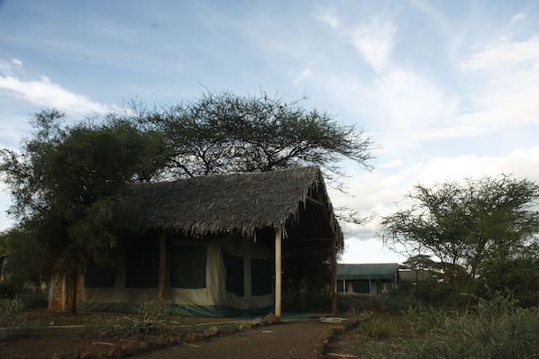 A thatched-roof structure with a covered entrance, surrounded by natural vegetation and trees. - Kimana Amboseli Camp (Amboseli)