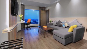 Een moderne woonkamer met een grijze bank, een houten salontafel, een aan de muur bevestigde televisie en een stadszicht door grote ramen.