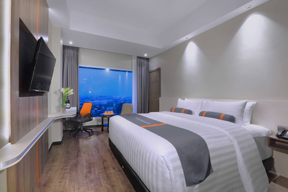 Habitación de hotel con una cama grande, un escritorio, una silla, un televisor y una ventana con vista a la ciudad.